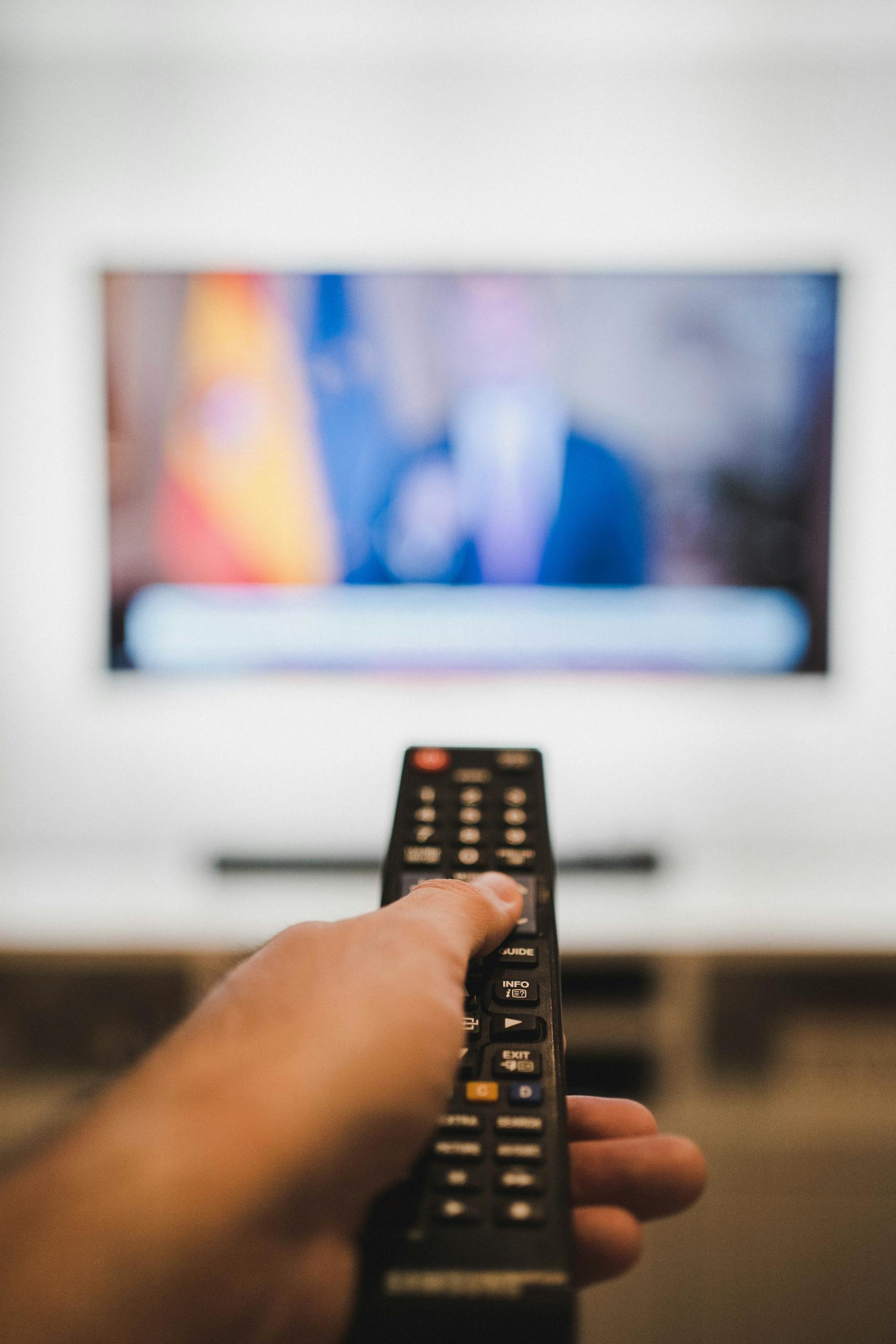 Close-up of a hand holding a remote control towards a TV screen indoors.